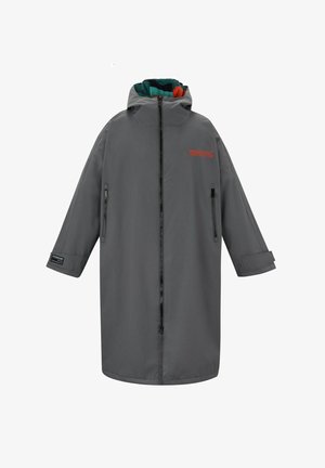 Hombre con un abrigo gris de gran tamaño con forro colorido, camisa naranja a rayas, pantalones cortos de mezclilla azules y zapatos marrones sin cordones, de pie contra un fondo sencillo.