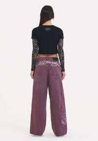 Svart kortärmad t-shirt över långärmad spetsblus, tillsammans med vida, blekta maroon byxor med broderad detalj och vit logotyp på baksidan.