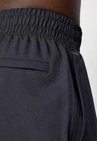 Mørkeblå joggebukser med synlig elastisk talje, glat tekstur, sidelomme og sydd detaljer langs sømmene.