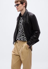 Μαύρο δερμάτινο μπουφάν με γιακά, φορεμένο πάνω από πουλόβερ με ασπρόμαυρο σχέδιο, σε συνδυασμό με μπεζ πλισέ παντελόνια.