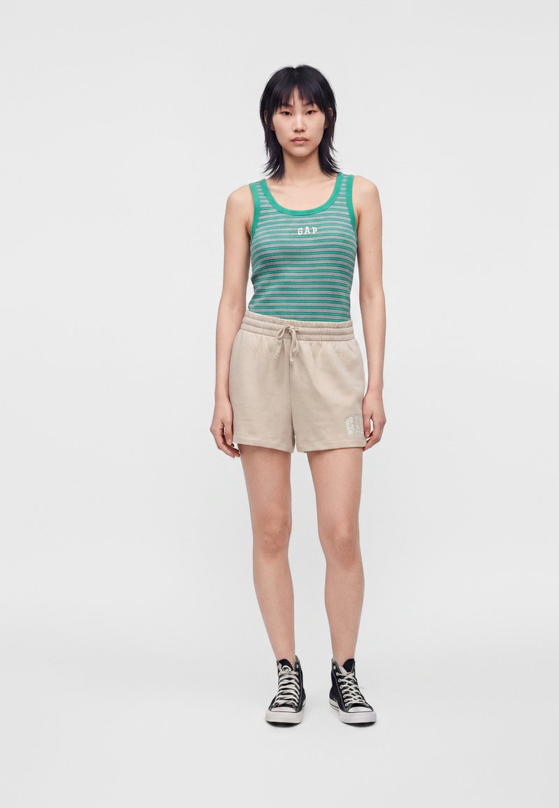 Femme debout portant un débardeur rayé vert et un short beige avec des baskets montantes noires sur un fond clair uni.