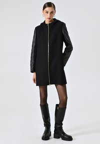 Cappotto nero con cappuccio e maniche in pelle nera trapuntata, dotato di zip frontale. Indossato con stivali neri fino al ginocchio e calze trasparenti.