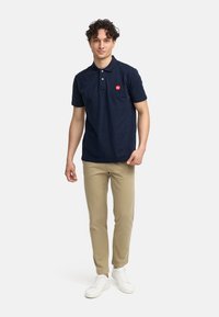 Mørkeblå polo med to knapper og en liten rød logo, kombinert med beige chinos og hvite sneakers. Enkelt og avslappet design.
