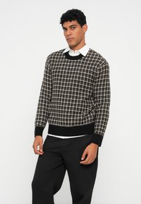 Άνδρας φορώντας καρό πουλόβερ σε μαύρο και λευκό, πάνω από άσπρο πουκάμισο με γιακά, φορώντας μαύρο παντελόνι, ποζάρει μπροστά σε απλό λευκό φόντο.