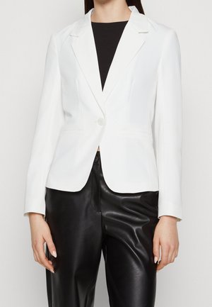Kvinde iført en tætsiddende hvid blazer med en knap og sorte læderbukser, stående mod en ensfarvet lys baggrund.