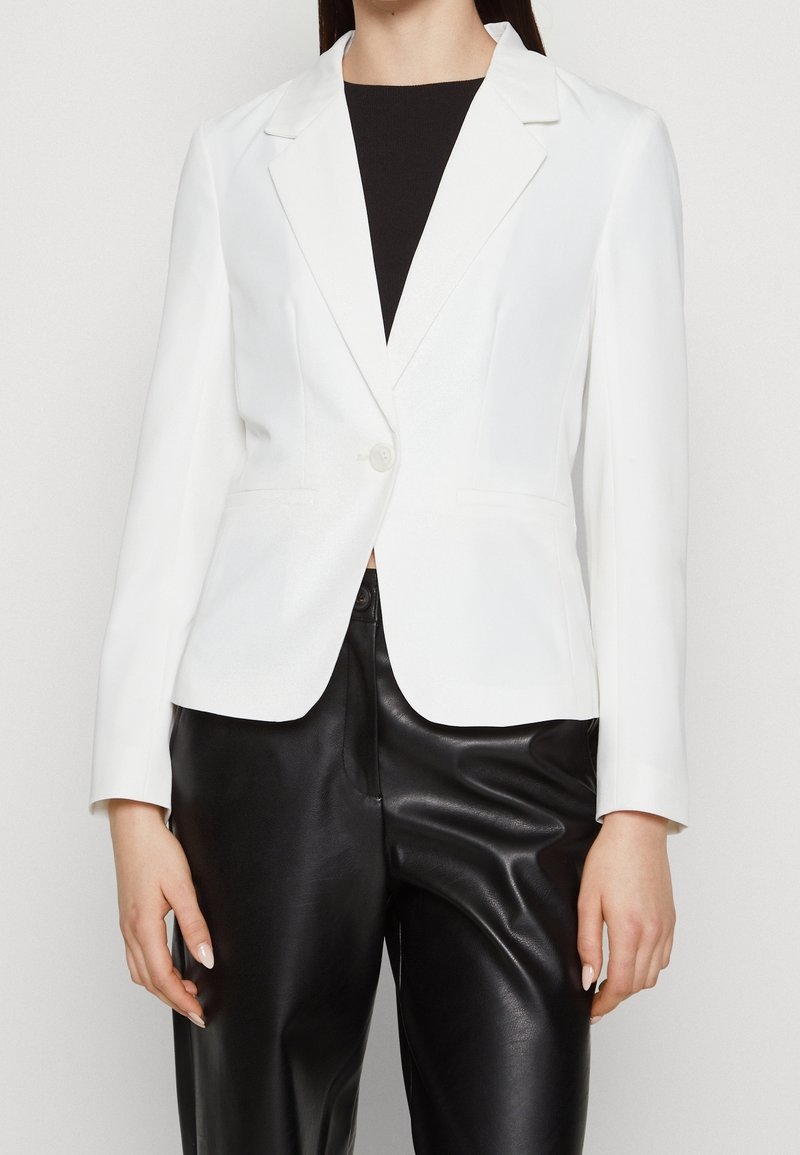 Femme portant un blazer blanc cintré avec un bouton et un pantalon en cuir noir, debout devant un fond clair uni.