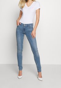 White short-sleeve V-neck T-shirt with a pink logo, paired with light blue skinny jeans and white high heels. Smooth fabric, fitted design.