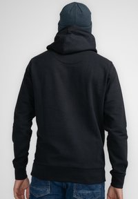 Sudadera negra con capucha ajustable, mangas largas y dobladillo acanalado, combinada con un gorro oscuro. La vista trasera muestra una textura de tela suave.