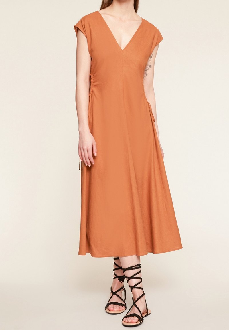 Robe couleur rouille avec un décolleté en V et des manches à emmanchures courtes, présentant une coupe fluide et des liens sur les côtés. Associée à des sandales à lanières noires.