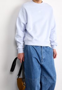 Light blue cotton sweatshirt with a crew neck, ribbed cuffs and hem, featuring a small white logo. Paired with loose denim pants and a brown bag.