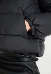 Doudoune noire en matériau brillant avec une poche zippée. Dispose de poignets élastiqués et d'un design légèrement rembourré.