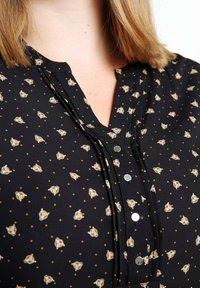 Gros plan sur un chemisier noir avec un petit motif tête de tigre et points, doté d'un devant plissé et de boutons-pression métalliques.