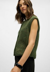 Pull sans manches en tricot vert avec une finition texturée, présentant un col rond et une coupe décontractée, associé à un pantalon noir et un bracelet noir.