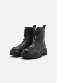 Botas de cuero negro hasta el tobillo con dos cremalleras laterales, una suela gruesa y una asa en bucle. Presentan una textura suave y un mínimo de costuras.