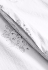 Biała tkanina z teksturowym, szarym haftem przedstawiającym kwiatowe wzory oraz ozdobnymi wycięciami. Gładka powierzchnia z guzikami wzdłuż krawędzi.