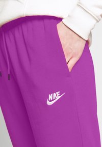 Violetti Nike-housut, joissa on nauhakuja, sivutaskut ja valkoinen logo. Valmistettu pehmeästä kankaasta, jossa on sileä pinta.