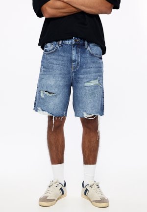 Homme portant un short en denim bleu déchiré, des chaussettes blanches et des baskets blanches avec des accents noirs, debout les bras croisés.