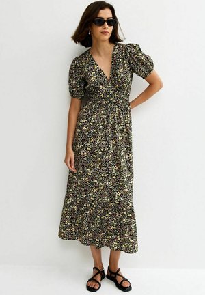 Mujer con vestido midi negro con flores y mangas abullonadas, sandalias negras de tiras y gafas de sol oscuras, de pie contra una pared blanca.