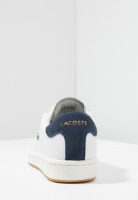 Baskets en cuir blanc avec un talon en daim bleu, comportant un logo brodé vert et une surface texturée. Lettrage doré sur le talon.