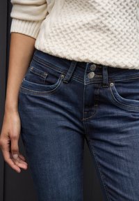 Mörkblå denimjeans har en figursydd design med framfickor, synliga sömmar och två metallknappar i midjan.