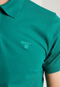 Zielona koszulka polo wykonana z teksturowanej bawełny. Posiada klasyczny kołnierzyk oraz haftowane logo na piersi. Krótkie rękawy z eleganckimi wykończeniami.
