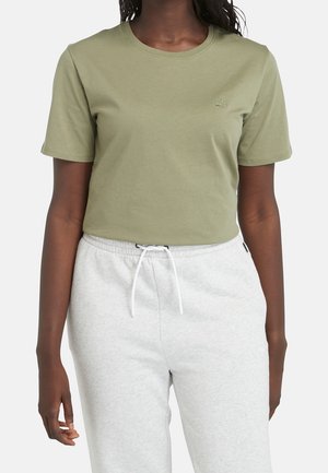 Vrouw die een licht olijfgroen T-shirt met korte mouwen en klein borstlogo draagt, en lichtgrijze joggingbroek met trekkoord tegen een effen achtergrond.