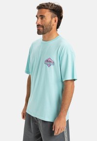 Light blue cotton t-shirt with a relaxed fit. Features a diamond-shaped graphic in pink and purple with "Quiksilver" text on the front.