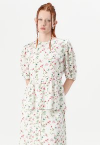Blusa bianca con maniche corte a sbuffo, dettagli a volant e un tessuto leggero e testurizzato con motivi floreali rosa e verdi.