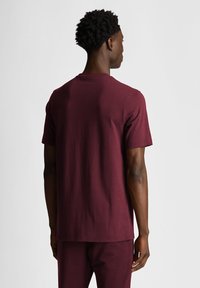 T-shirt in cotone bordeaux con maniche corte, scollo rotondo e vestibilità ampia, visto da dietro. Texture liscia senza fantasie.