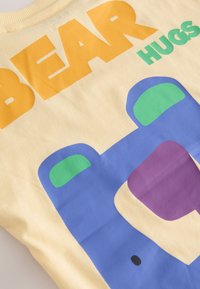 Yellow cotton t-shirt featuring large orange text "BEAR HUGS" and colorful bear graphic in blue, green, and purple with smooth texture.