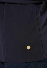 Diepblauwe top van een gladde stof, met een decoratieve goudkleurige logo-accent en subtiele stikdetails langs de zoom.