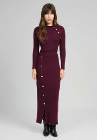 Femme portant une robe bordeaux à manches longues et longue jusqu'aux chevilles, avec des boutons argentés sur le côté et à l'avant, assortie à des bottes à talons noires.