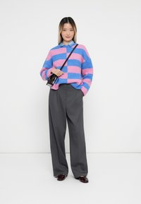 Ženska v roza in modri črtasti puloverju, sivih širokih hlačah, temnih čevljih in črni torbi čez ramo na belem ozadju.