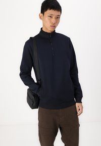 Felpa con zip navy e collo alto, con polsini e orlo a coste, abbinata a pantaloni cargo marroni. Una borsa a tracolla nera completa il look.
