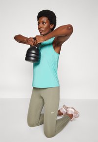 Una pesa rusa negra es sostenida por una persona que lleva una camiseta sin mangas azul claro y leggings oliva, con zapatos rosa claro. Fondo simple.