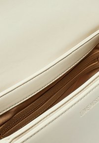 Interno della borsa color crema con una texture liscia. Presenta una tasca con zip marrone e un logo impresso lungo il bordo.