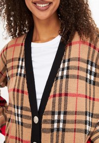 Cárdigan de cuadros beige con acentos negros, que presenta un escote en V profundo, botones plateados y una mezcla de patrones de cuadros en rojo, negro y blanco.
