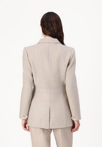 Beige herringbone blazer med en skreddersydd passform, støtteklaffer og belted midje. Har knapper på mansjettene og en splitt bak.
