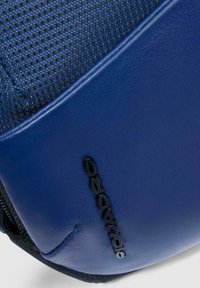 Borsa da viaggio blu con superficie in tessuto texturizzato, con una sezione liscia simile alla pelle e un accento logo nero. Dettaglio della cerniera visibile.