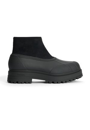 Mustat nilkkurit, joissa on teksturoitu kumista ja mokasta valmistettu yläosa, pyöristetty kärki ja paksu, uritettu pohja pitävää otetta varten.