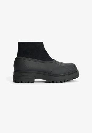 Mustat nilkkurit, joissa on teksturoitu kumista ja mokasta valmistettu yläosa, pyöristetty kärki ja paksu, uritettu pohja pitävää otetta varten.