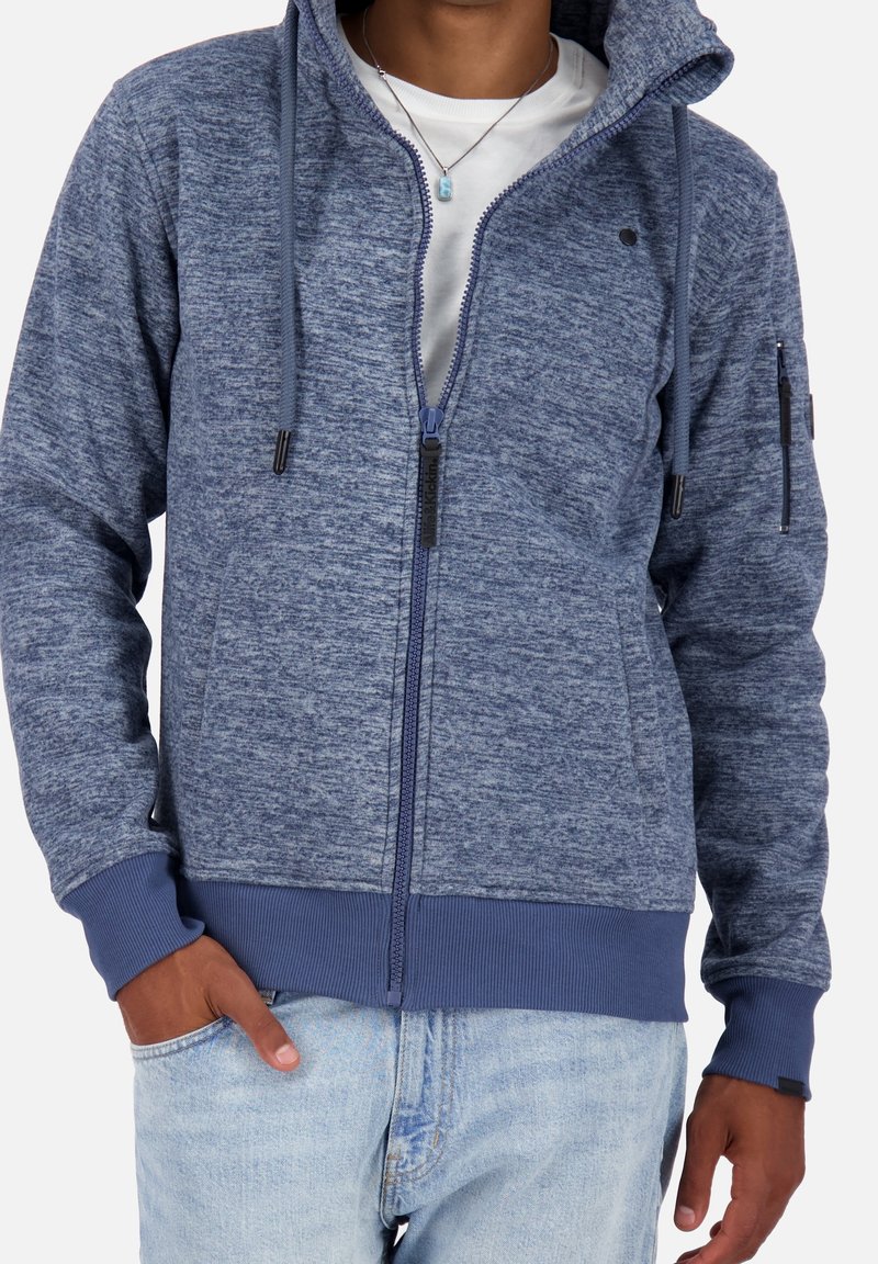 Mężczyzna ubrany w niebieską rozpinaną bluzę z kapturem ze ściągaczami, białą koszulkę, jasnoniebieskie dżinsy i naszyjnik z zawieszką.
