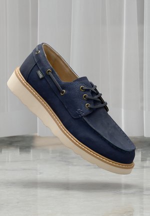 Mørkeblå sejlersko i ruskind med lys tan-sål, læderfor, messingøjer og sorte snørebånd, der svæver over en marmeroverflade.