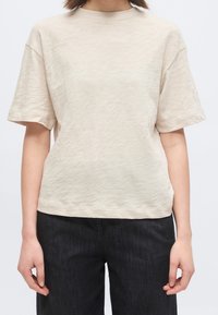 Personne portant une chemise à manches courtes beige clair et un pantalon foncé, montrée des épaules aux hanches sur un fond uni.