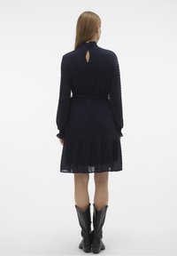 Mörkblå klänning med texturerade prickar, långa ärmar, b älte vid midjan och volang i nederkant. Har en nyckelhålsdetalj i ryggen. Bärs med svarta stövlar.