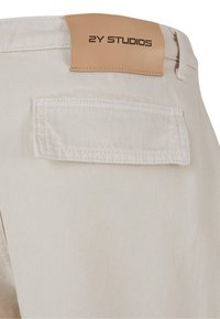Pantalon en denim beige à surface texturée, avec une poche arrière et une étiquette en cuir marron avec l'inscription « 2Y STUDIOS » embossée.