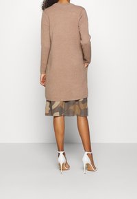 Cardigan en tricot à manches longues, de couleur marron clair, avec une coupe décontractée. Porté par-dessus une jupe à volants à motif camouflage et des chaussures à talons blancs.