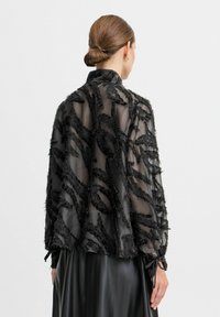 Blusa nera trasparente con un motivo testurizzato di fili neri, maniche lunghe con lacci e una vestibilità ampia. Il tessuto è leggero e fluido.