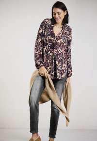 Langärmlige, florale Bluse in burgunderroten und cremefarbenen Mustern, kombiniert mit lässigen grauen Jeans. Ein beiger Pullover über den Armen drapiert.