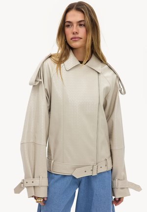Femme portant une veste beige texturée avec un col large, des poignets ceinturés et une ceinture assortie, associée à un jean bleu, regardant légèrement vers la droite.